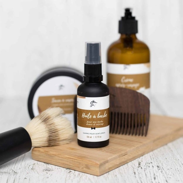Flacon d’huile à barbe naturelle pour homme avec bouchon en sprayc, sur fond neutre, entouré de brosse et peigne sur bois de cèdre.