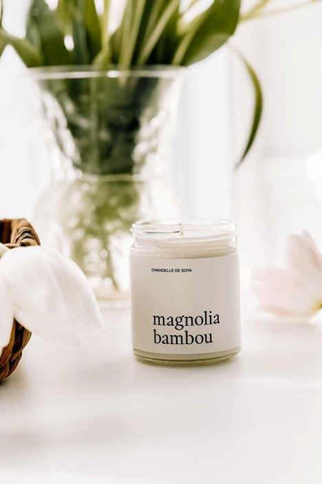 Chandelle de soya Magnolia Bambou dans un pot en verre, posée sur un comptoir blanc avec des tiges de bambou en arrière-plan, créant une ambiance zen et naturelle.