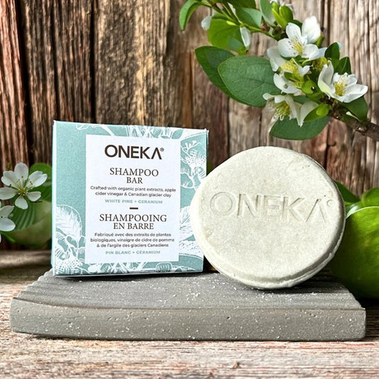 Shampoing en barre naturel au pin blanc et géranium sur un porte-savon , entouré d’ingrédients botaniques comme des feuilles de pin, fleurs de géranium et argile.